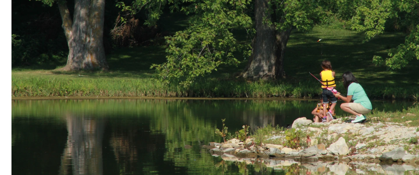 Two children in life jackets fishing with their mom on a small rock outcropping on a lake.